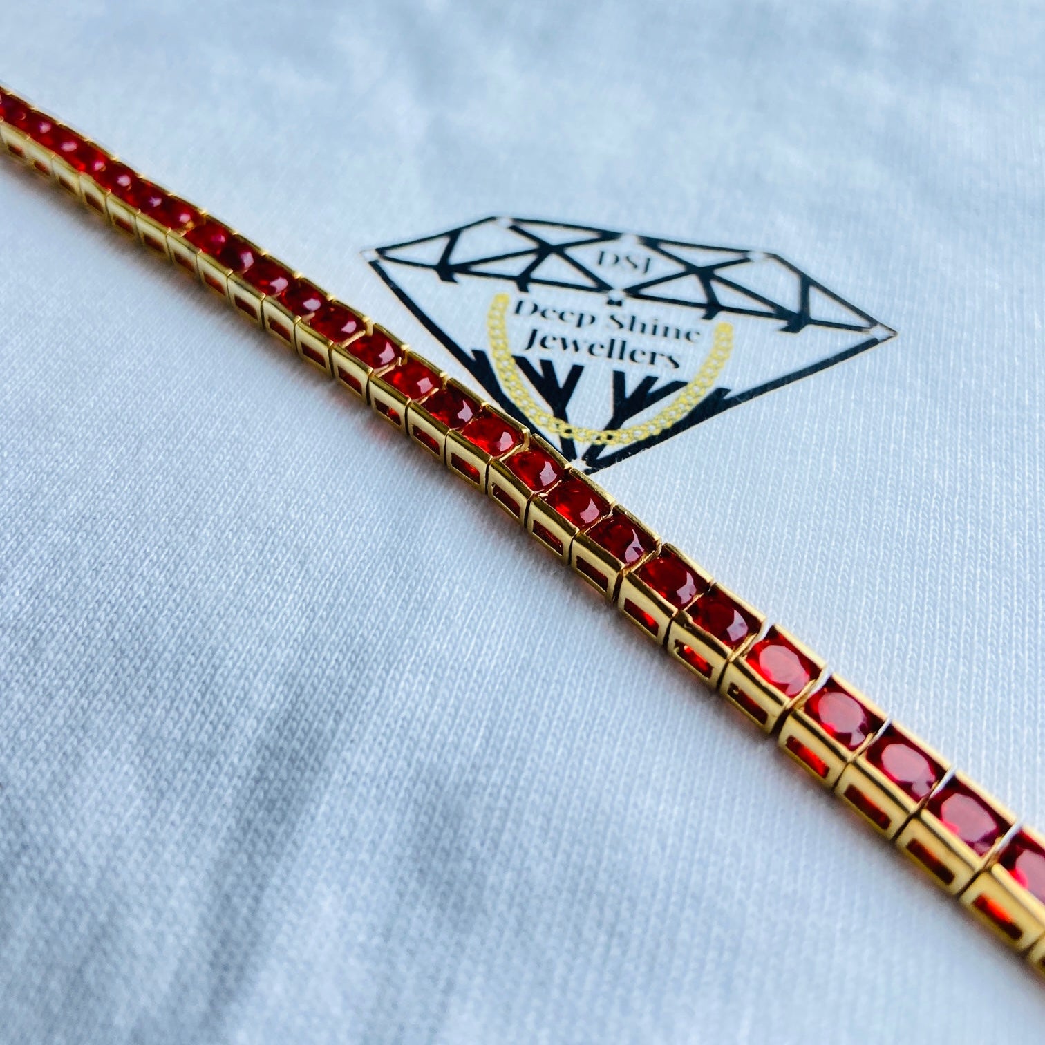 Gold bracelet with red stones on a white shirt with a brand logo. Gold bracelet with red gemstones on a white cushion Gold red ruby bracelet women queens ladies female tennis bracelet ruby red 925 silver sterling silver gold plated golden exclusive affordable luxury jewellery jewelry Deep Shine Jewellers DSJ Your Time To Shine trending best seller buy purchase bracelets 