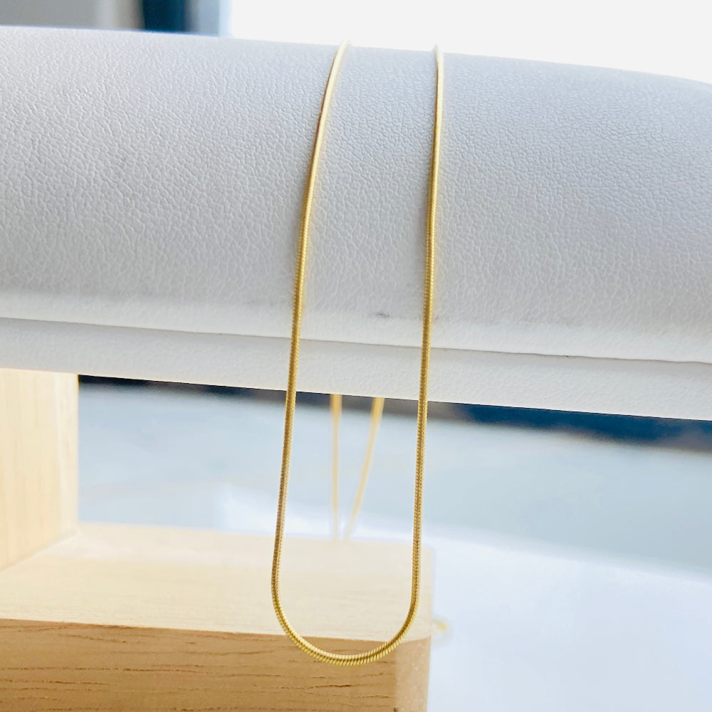 Gold necklace on a white surface with a wooden stand Gold bar on a light blue marble surface gold snake bone chain women men ladies gents kinds queens exclusive craftsmanship affordable luxury high quality premium jewellery jewelry Deep Shine Jewellers gold plated golden 925 silver sterling silver Your Time To Shine DSJ