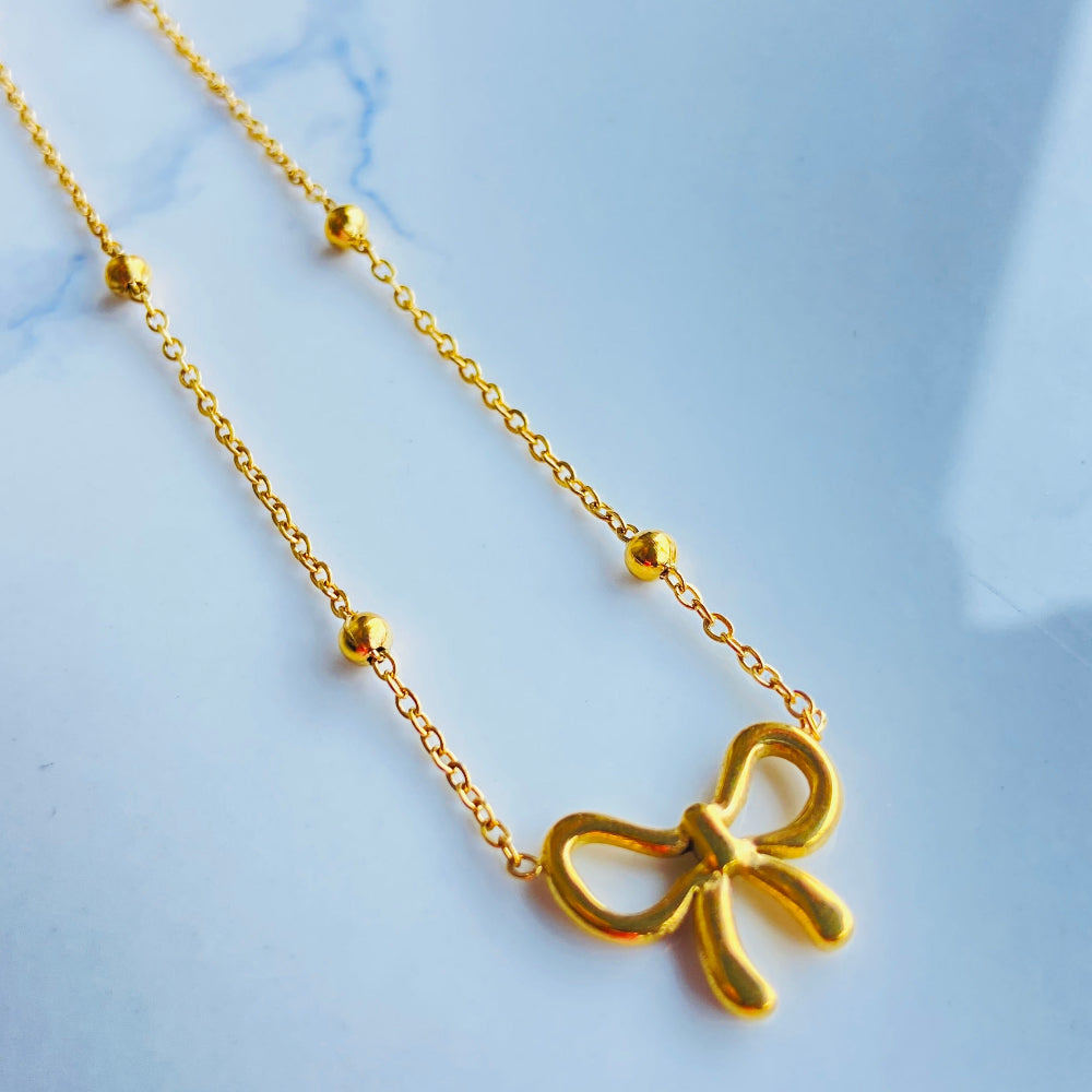 Gold bow-shaped necklace on a light blue marble background old bow-shaped pendant on a chain against a light blue background gold bow balled chain necklace gold plated golden women ladies queens stainless steel premium jewellery jewelry affordable luxury Deep Shine Jewellery Your Time To Shine best sellers trending for him her
