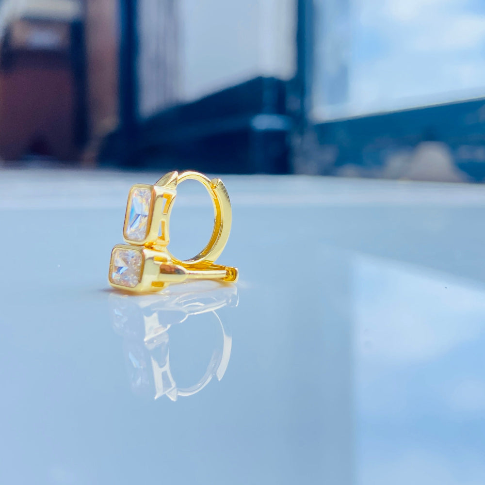 Gold ring with a clear gemstone on a reflective surface Gold hoop earring with clear stones on a reflective surface with blue sky background gold single stone sleeper earrings women ladies queens gold plated golden cubic zirconia diamond baguette 925 silver sterling silver exclusive craftsmanship high quality premium affordable luxury Deep Shine Jewellers jewellery jewelry Your Time To Shine DSJ