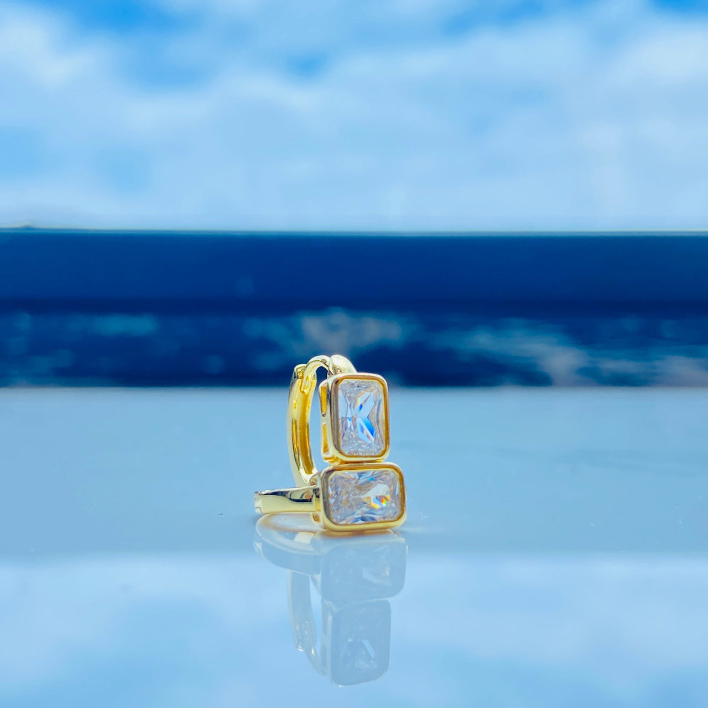 Gold ring with a blue gemstone on a reflective surface with a blue sky background Gold hoop earring with clear stones on a reflective surface with blue sky background gold single stone sleeper earrings women ladies queens gold plated golden cubic zirconia diamond baguette 925 silver sterling silver exclusive craftsmanship high quality premium affordable luxury Deep Shine Jewellers jewellery jewelry Your Time To Shine DSJ