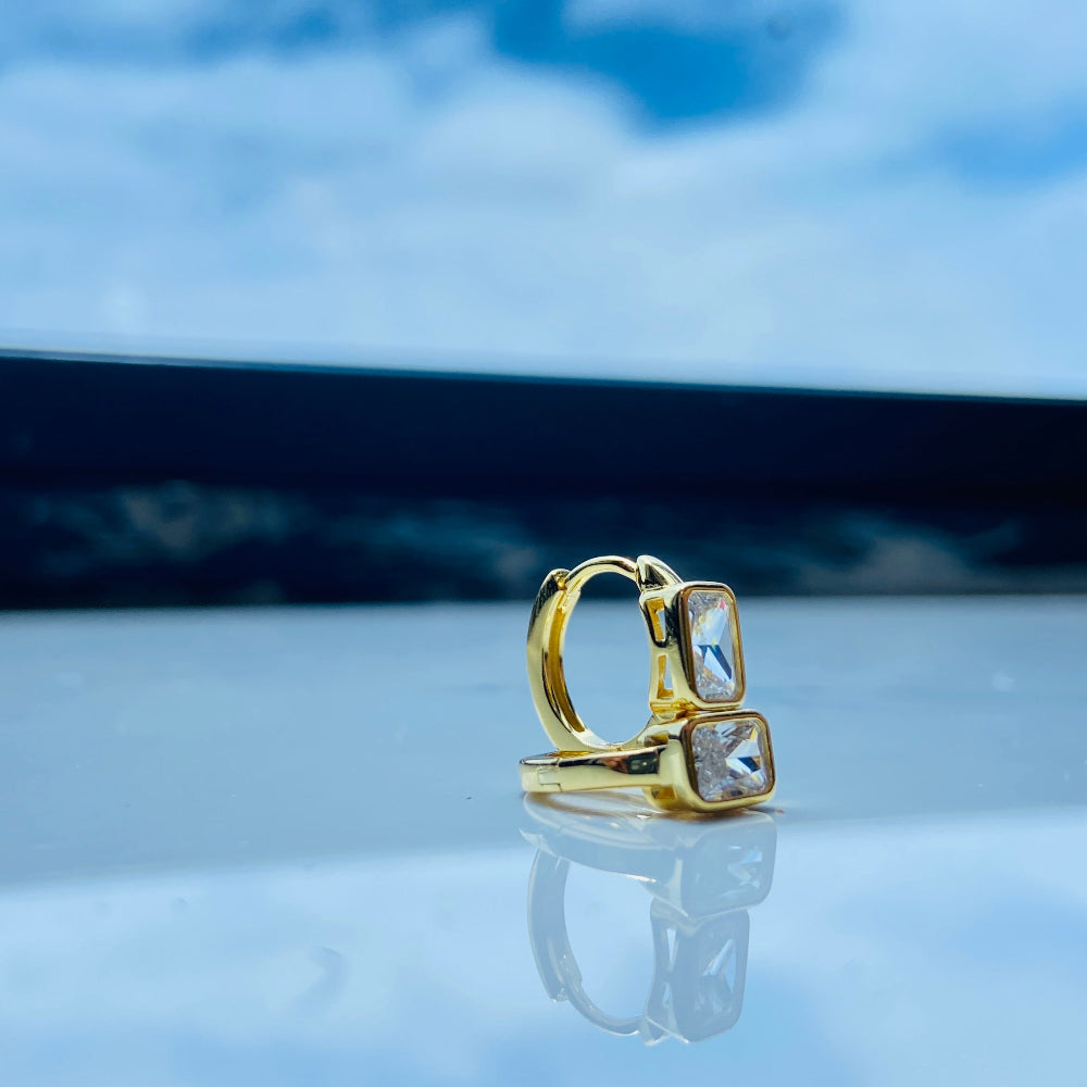 Gold hoop earring with clear stones on a reflective surface with blue sky background gold single stone sleeper earrings women ladies queens gold plated golden cubic zirconia diamond baguette 925 silver sterling silver exclusive craftsmanship high quality premium affordable luxury Deep Shine Jewellers jewellery jewelry Your Time To Shine DSJ
