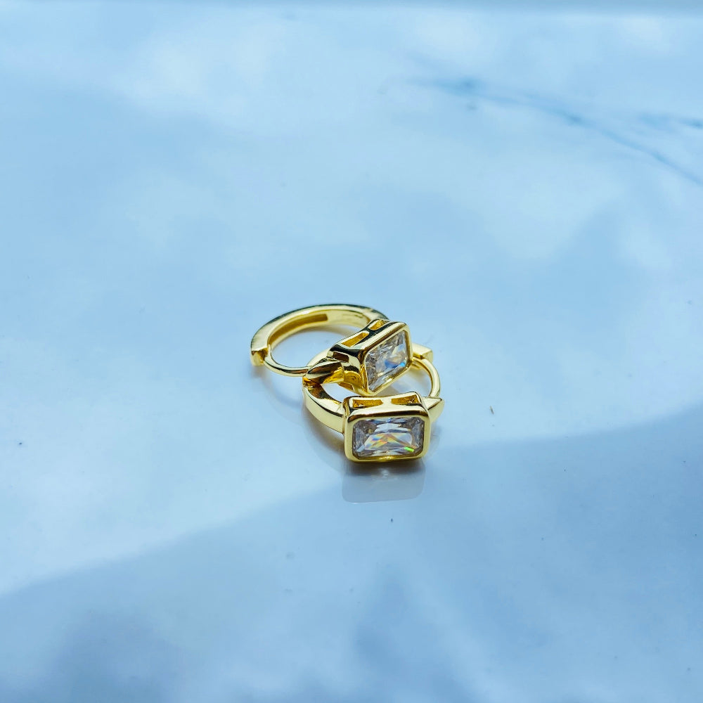 Gold earring with clear stones on a light blue fabric background Gold hoop earring with clear stones on a reflective surface with blue sky background gold single stone sleeper earrings women ladies queens gold plated golden cubic zirconia diamond baguette 925 silver sterling silver exclusive craftsmanship high quality premium affordable luxury Deep Shine Jewellers jewellery jewelry Your Time To Shine DSJ