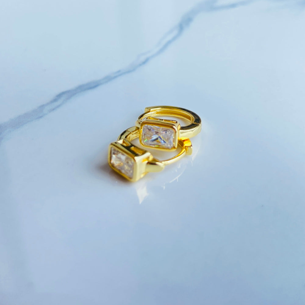 Gold hoop earring with clear stones on a reflective surface with blue sky background gold single stone sleeper earrings women ladies queens gold plated golden cubic zirconia diamond baguette 925 silver sterling silver exclusive craftsmanship high quality premium affordable luxury Deep Shine Jewellers jewellery jewelry Your Time To Shine DSJ