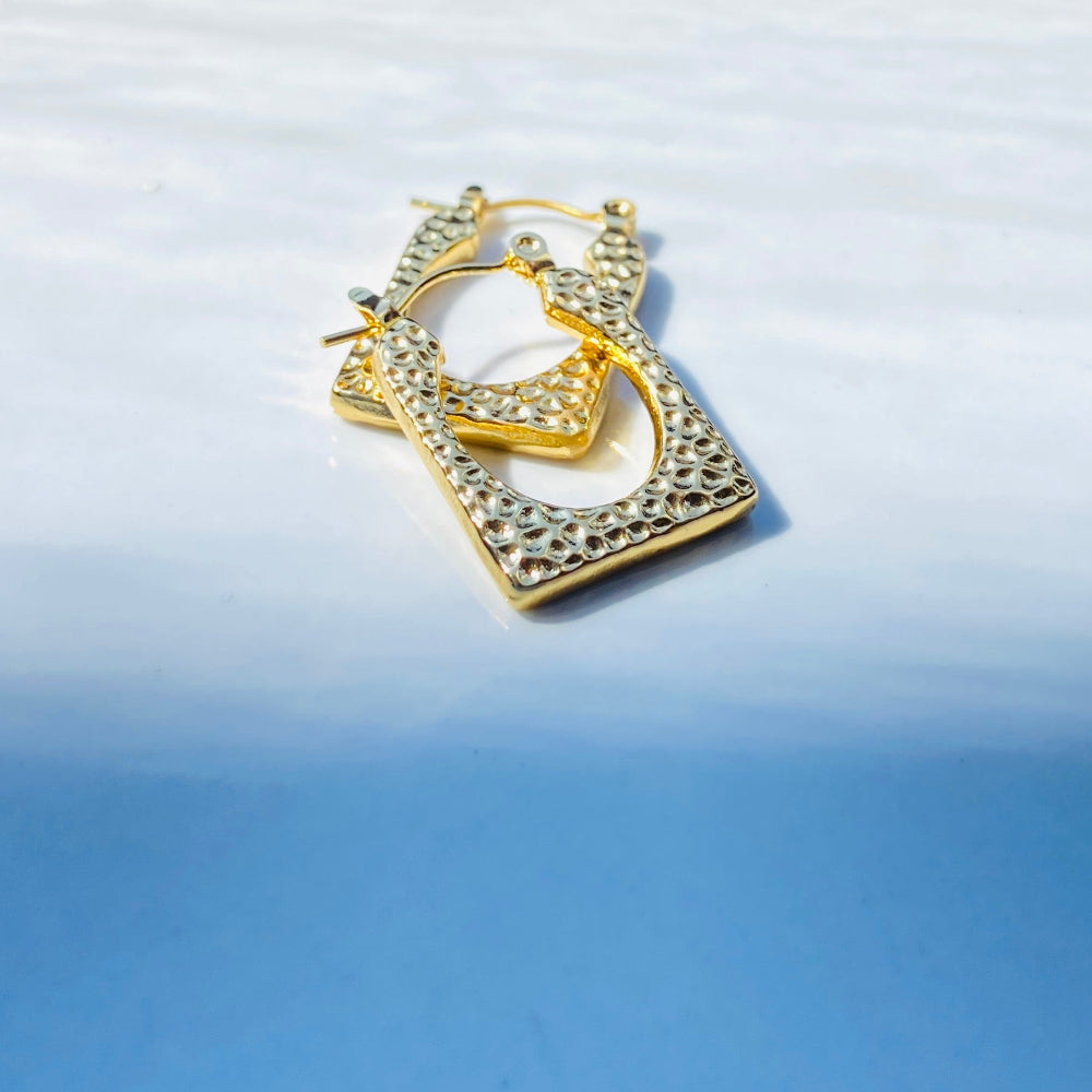 Gold heart-shaped earring on a reflective surface Gold hoop earrings on a light blue surface gold detailed rectangle earrings exclusive by Deep Shine Jewellers jewellery jewelry DSJ Your Time To Shine golden shine